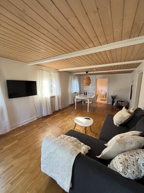 A living room with a wooden ceiling, a flat-screen TV mounted on the wall, a dark sofa with patterned cushions, a white round table with chairs, and a small white stool. - Charming house with beautiful views (Gedser)