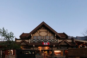 A building with a prominent Canadian flag, storefronts labeled 'sevenfold' and 'Ken Hoehn Gallery,' and a mountainous backdrop. - Sevenfold | Sleeps 20 | Luxe 7BR Retreat + Hot Tub (Canmore)