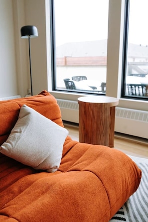 A room with a large window, a sofa with a pillow, a floor lamp, and a small wooden table.