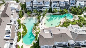 Resort com vários edifícios, piscina central e palmeiras ao redor.