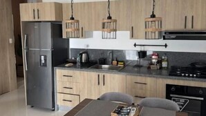 Une cuisine moderne avec des meubles en bois, un réfrigérateur en acier inoxydable, une table à manger avec deux chaises et des luminaires suspendus.