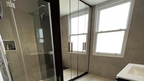 A modern bathroom with a glass shower enclosure, a walk-in closet with mirrored doors, and a large window providing natural light. - Luxurious Fulham Apartment (London)