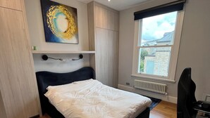 A bedroom with a bed, a desk, a chair, a radiator, a window, and a ceiling light fixture. - Luxurious Fulham Apartment (London)