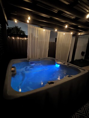 A hot tub with a blue light, surrounded by a wooden deck and white curtains.