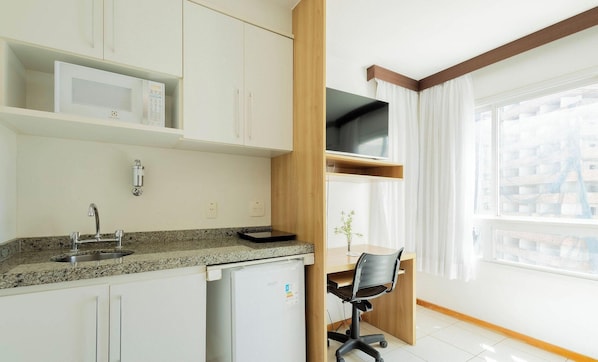 Cozinha compacta com micro-ondas embutido, pia e pequena geladeira. Há uma escrivaninha de madeira com cadeira e uma televisão montada acima. O quarto possui uma janela grande com cortinas.