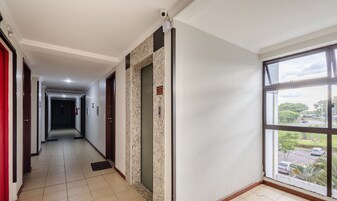 Corredor de hotel com piso de azulejos, detector de incêndio, câmera de segurança e janela com vista para um parque de estacionamento e árvores.