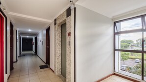 A hotel corridor with a tiled floor, a fire alarm, a security camera, and a window with a view of a parking lot and trees. - Comfortable Apartment, Lago Norte Bvt0418 (Lago Norte)