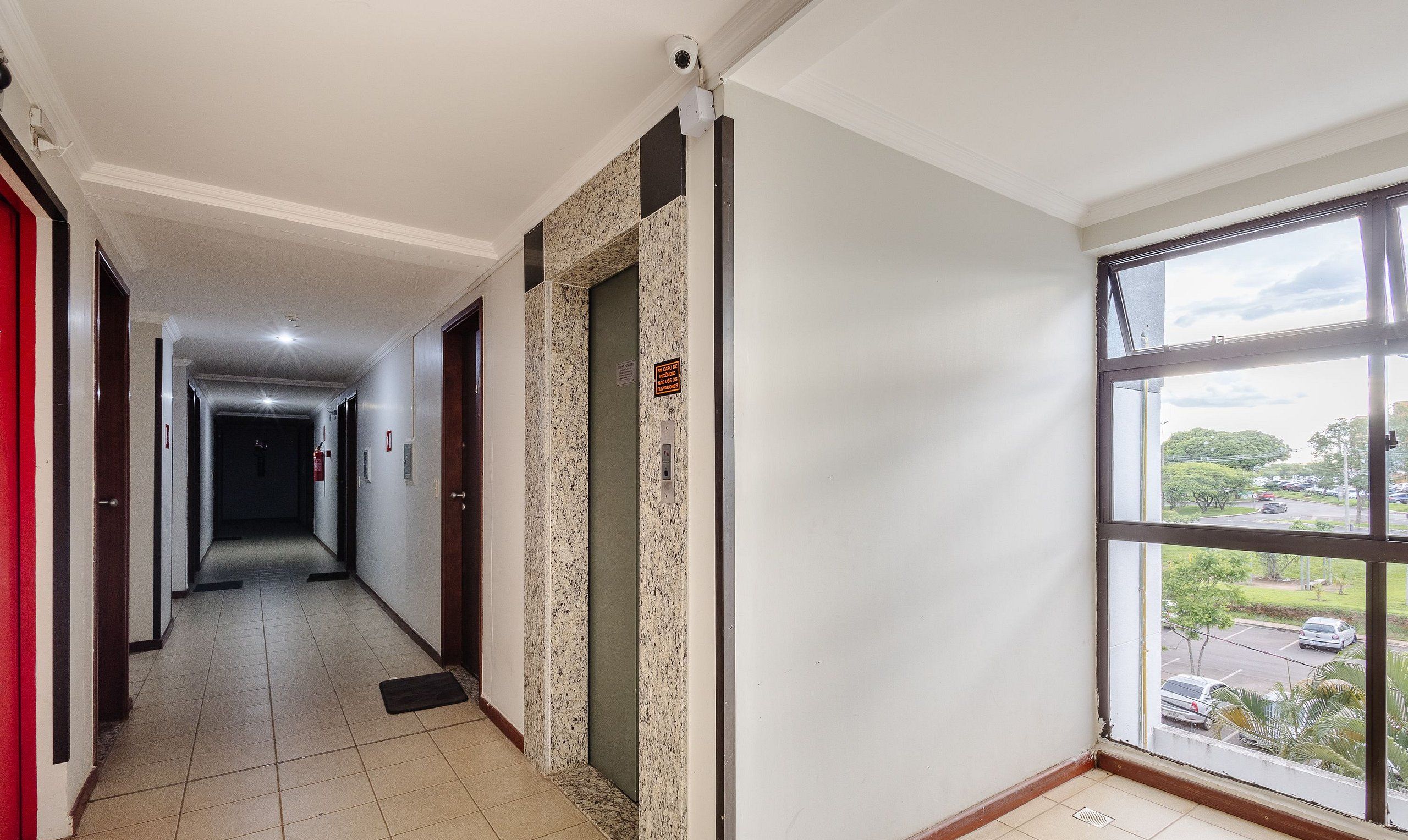 Un couloir d’hôtel avec un sol carrelé, un détecteur d’incendie, une caméra de surveillance et une fenêtre donnant sur un parking et des arbres.