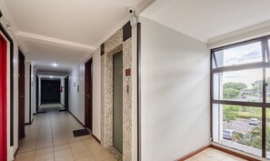A hotel corridor with a tiled floor, a fire alarm, a security camera, and a window with a view of a parking lot and trees. - Comfortable Apartment, Lago Norte Bvt0418 (Lago Norte)