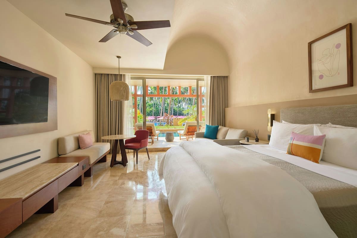 A hotel room with a large bed, a ceiling fan, a TV mounted on the wall, a seating area with a table and chairs, and a view of the outdoors.