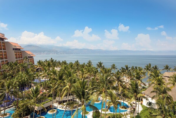 A resort with multiple swimming pools surrounded by palm trees, a view of the ocean, and a mountain range in the distance.
