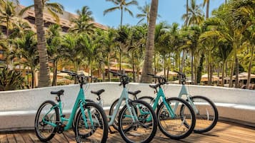 A row of turquoise electric bicycles parked on a wooden deck with palm trees and a building in the background.