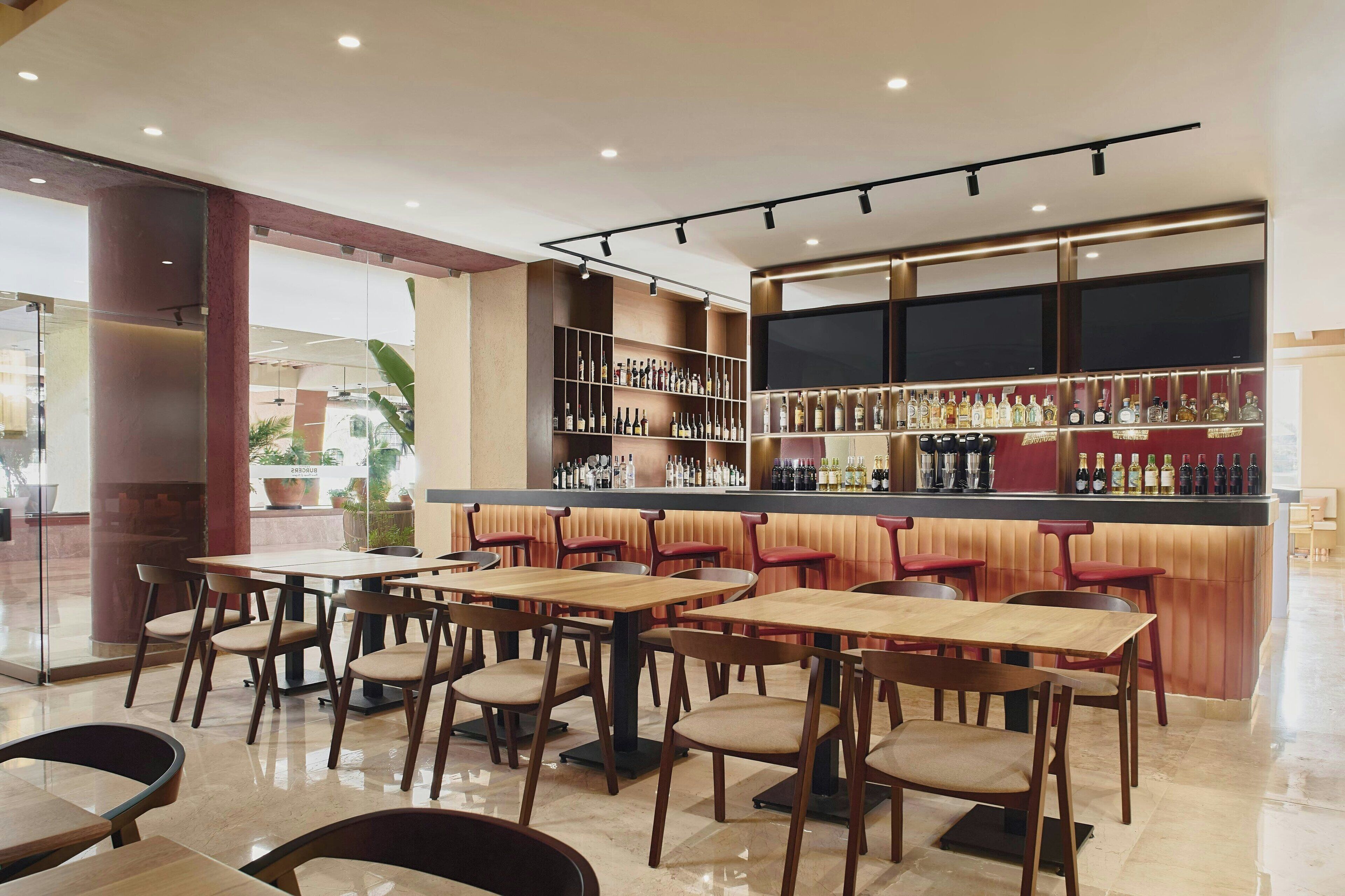 A modern bar area with wooden tables and chairs, a well-stocked liquor shelf, and a large TV screen.