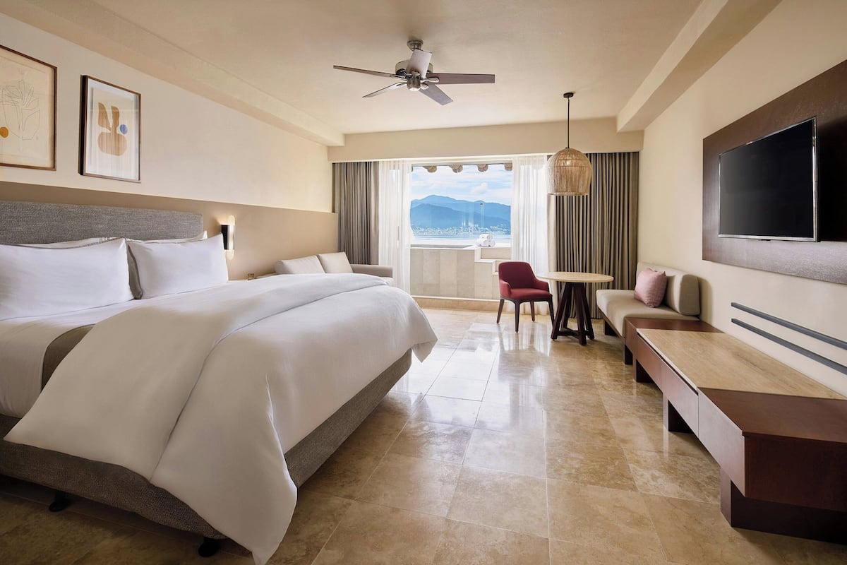 A hotel room with a large bed, a desk, a sofa, a TV, and a balcony with a view.
