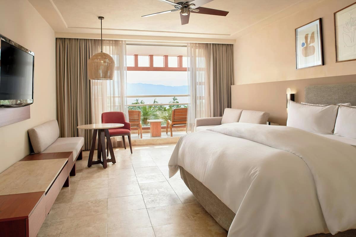 A hotel room with a large bed, a TV mounted on the wall, a ceiling fan, a balcony with a view of the mountains, and a dining area with a table and chairs.