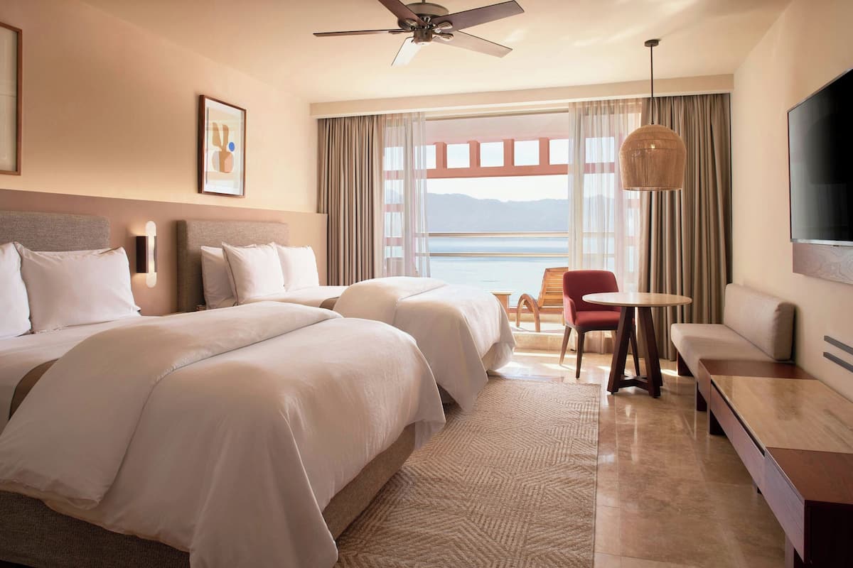 A hotel room with two beds, a ceiling fan, a TV, a sofa, a table, and a view of the sea.