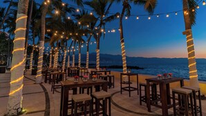 An outdoor seating area with tables and chairs, decorated with string lights and palm trees, overlooking a body of water. - The Westin Playa Vallarta, An All-Inclusive Resort (Puerto Vallarta)