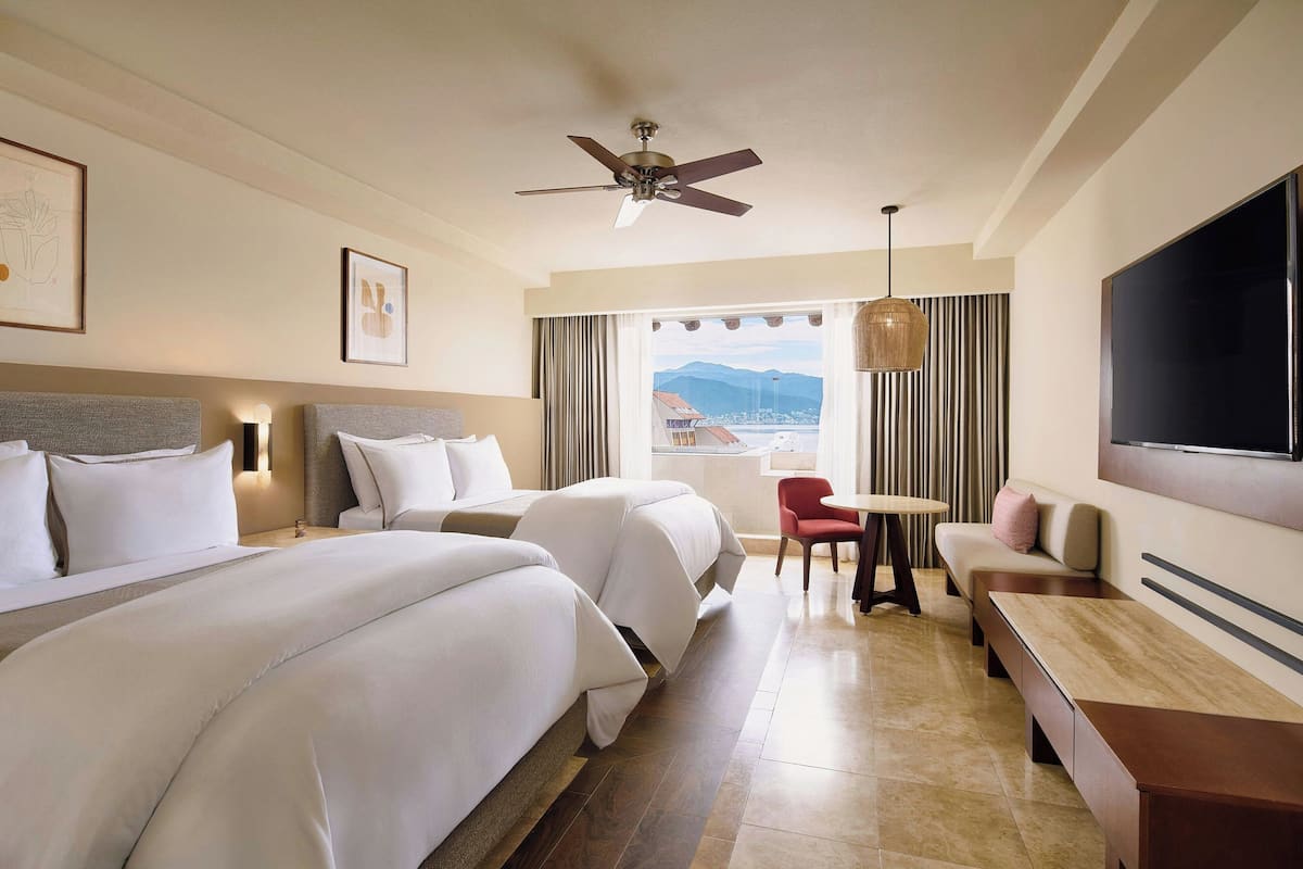 A hotel room with two beds, a ceiling fan, a TV, a dining table, and a view of mountains.
