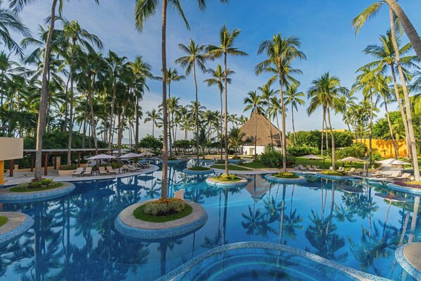 A resort pool area with multiple circular water features surrounded by palm trees and lounge chairs.