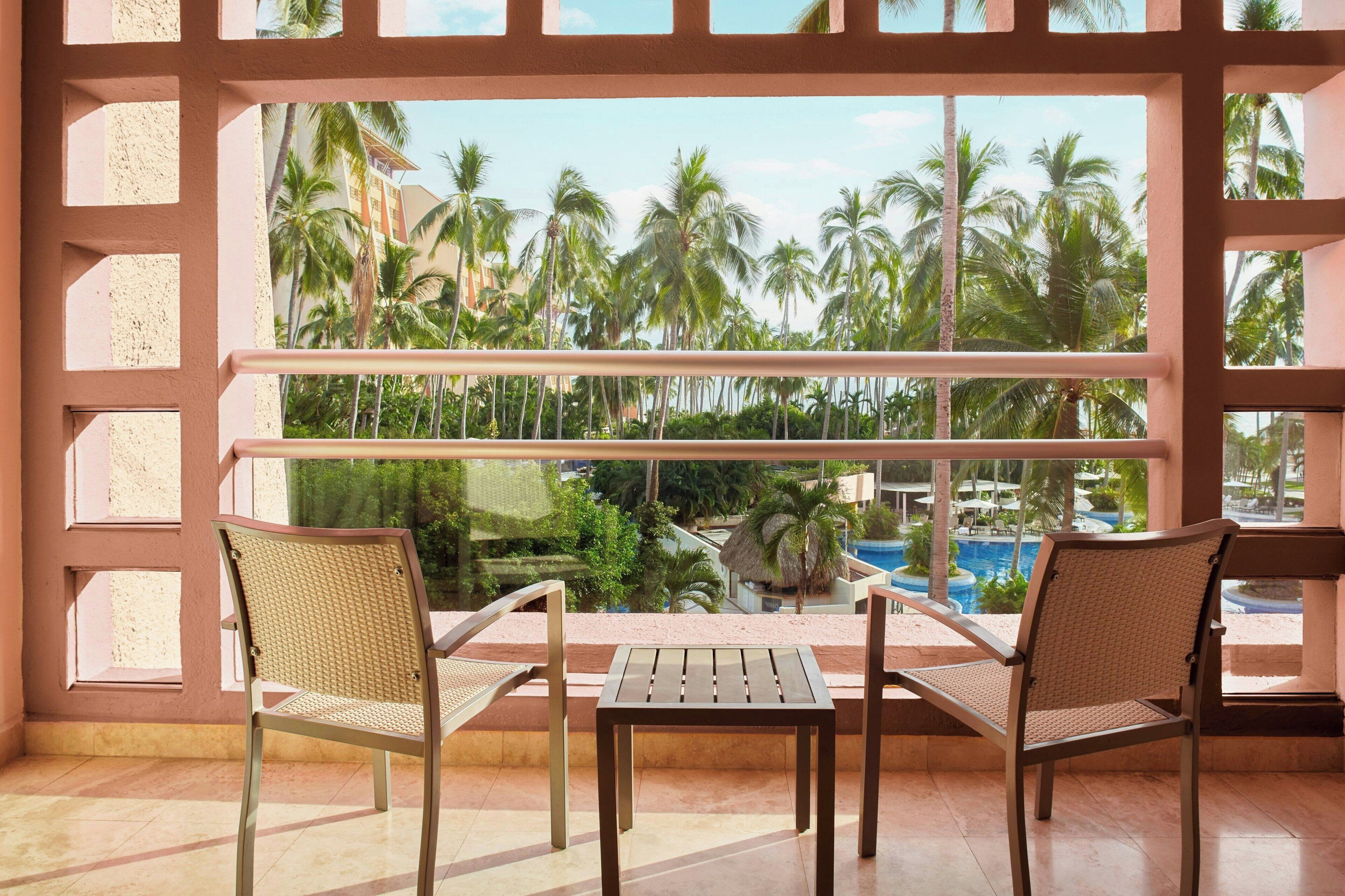 Un balcon avec deux fauteuils en osier et une petite table, donnant sur une piscine et des palmiers.