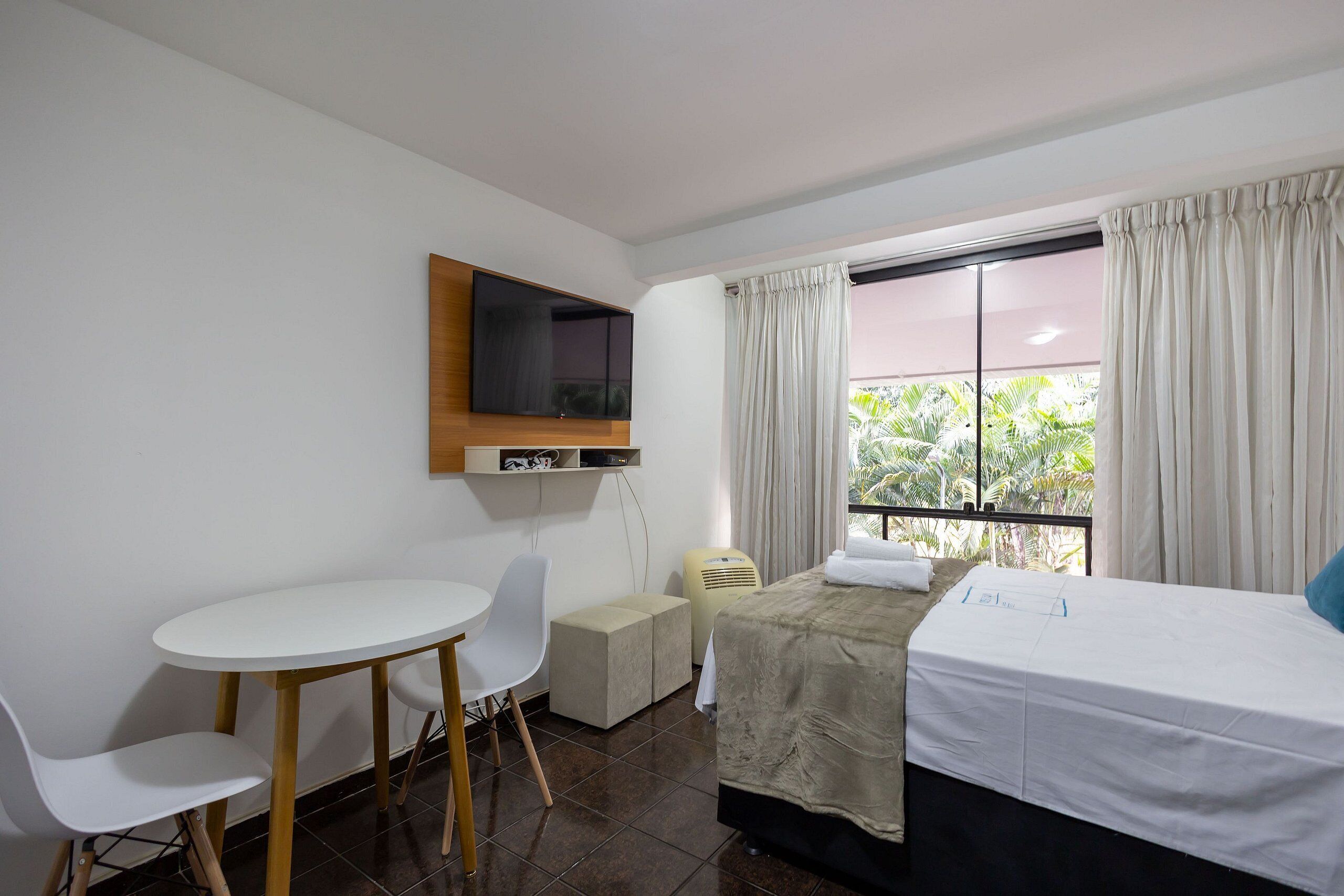 Habitación de hotel con cama, escritorio y sillas, televisor montado en la pared y ventana con cortinas.
