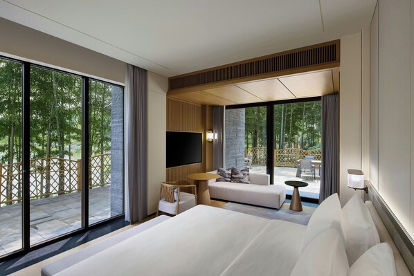 A modern hotel room with a large bed, a sofa, a TV, and a balcony with a view of trees.