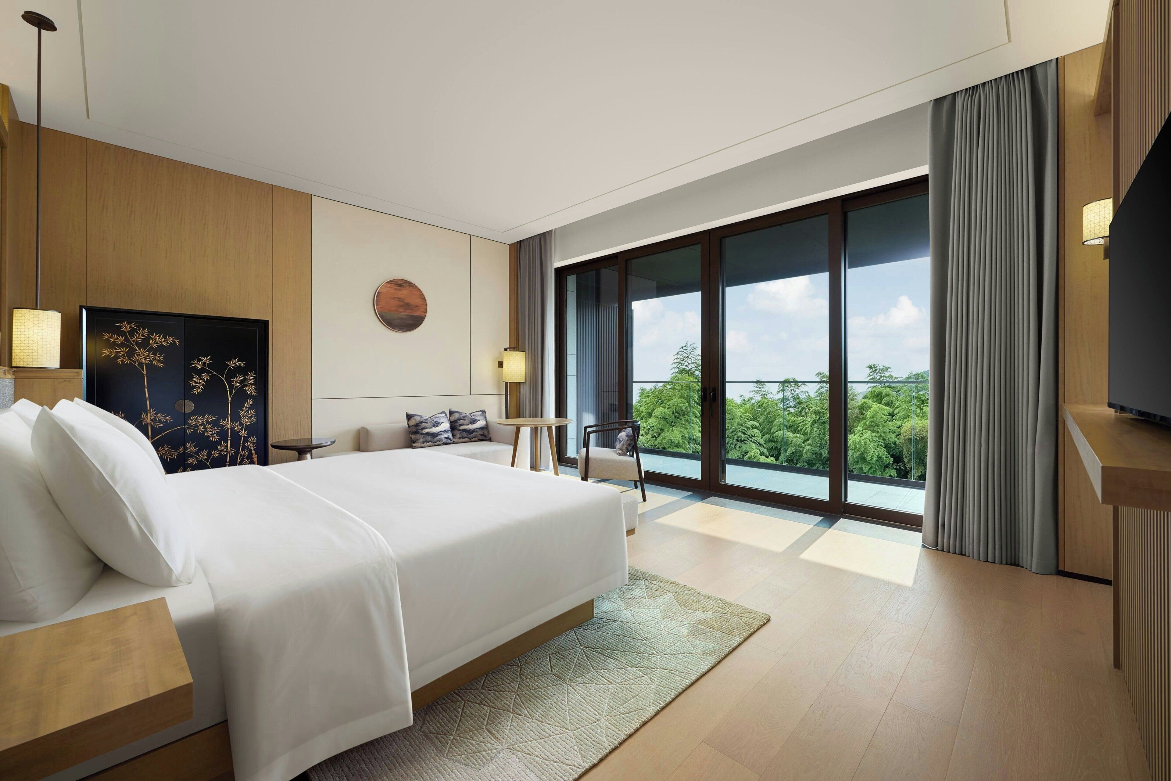 Ein modernes Hotelzimmer mit einem großen Bett, Blick ins Grüne durch die Fenster und einem kleinen Tisch mit Stuhl.