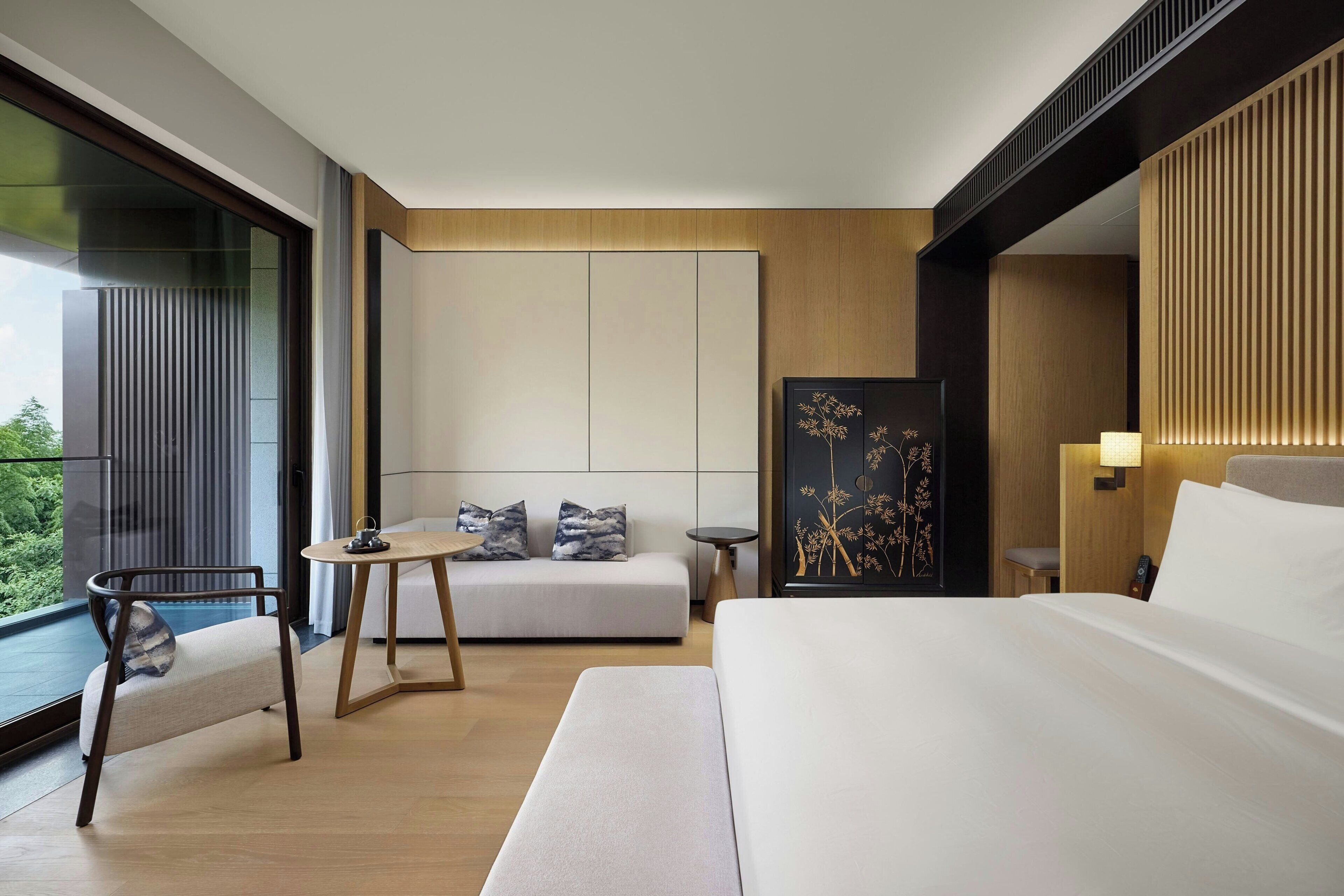 Camera d'albergo moderna con un letto grande, una poltrona, un tavolino e una sedia. Un'ampia finestra offre vista su una zona verde.