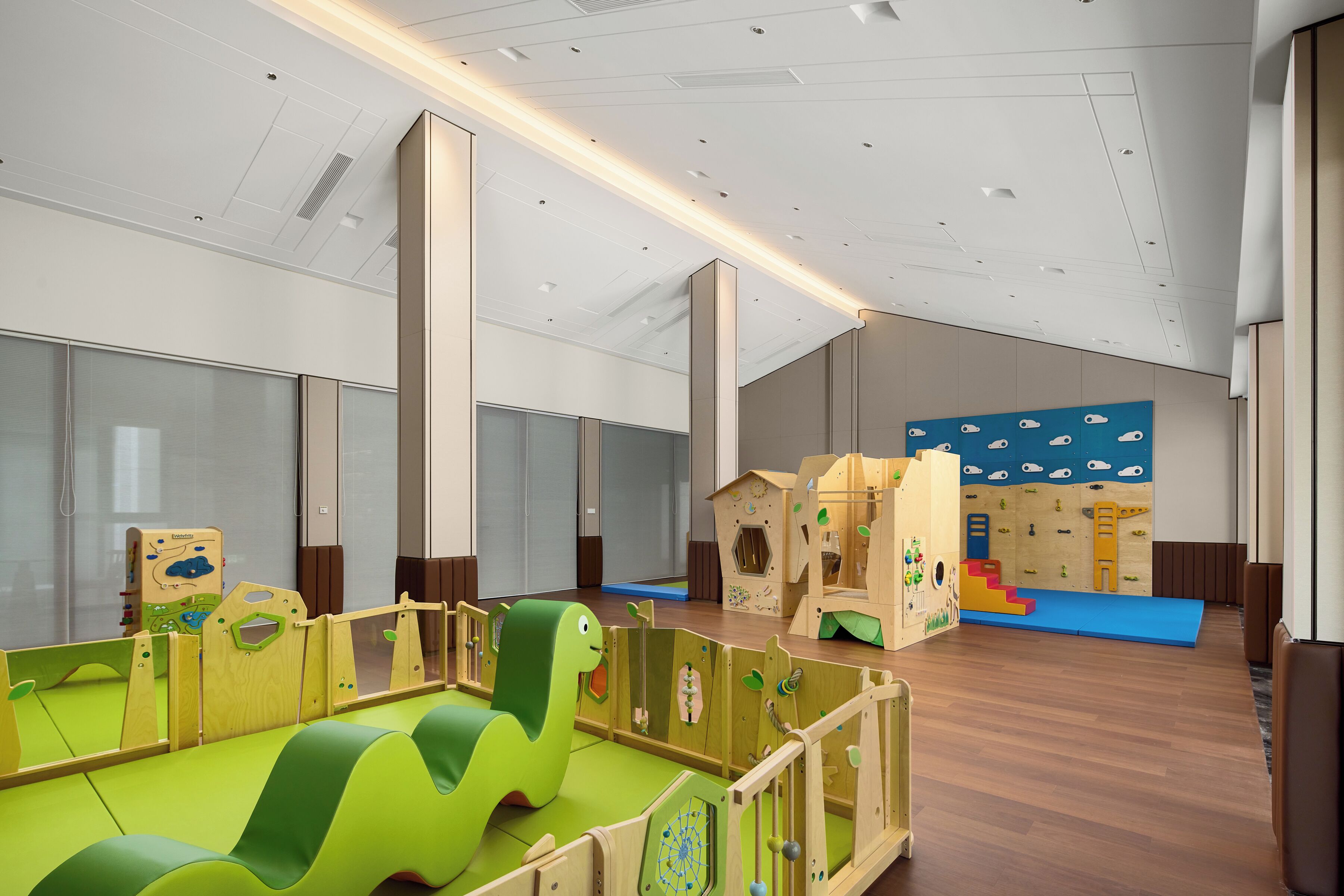 A spacious play area with soft play mats, wooden play structures, and a large window providing natural light.