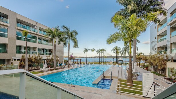 A swimming pool with a view of the sea, surrounded by palm trees and modern buildings.