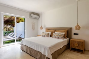Quarto com uma cama grande, uma mesa de cabeceira de madeira e vista para área externa com piscina e espreguiçadeiras.
