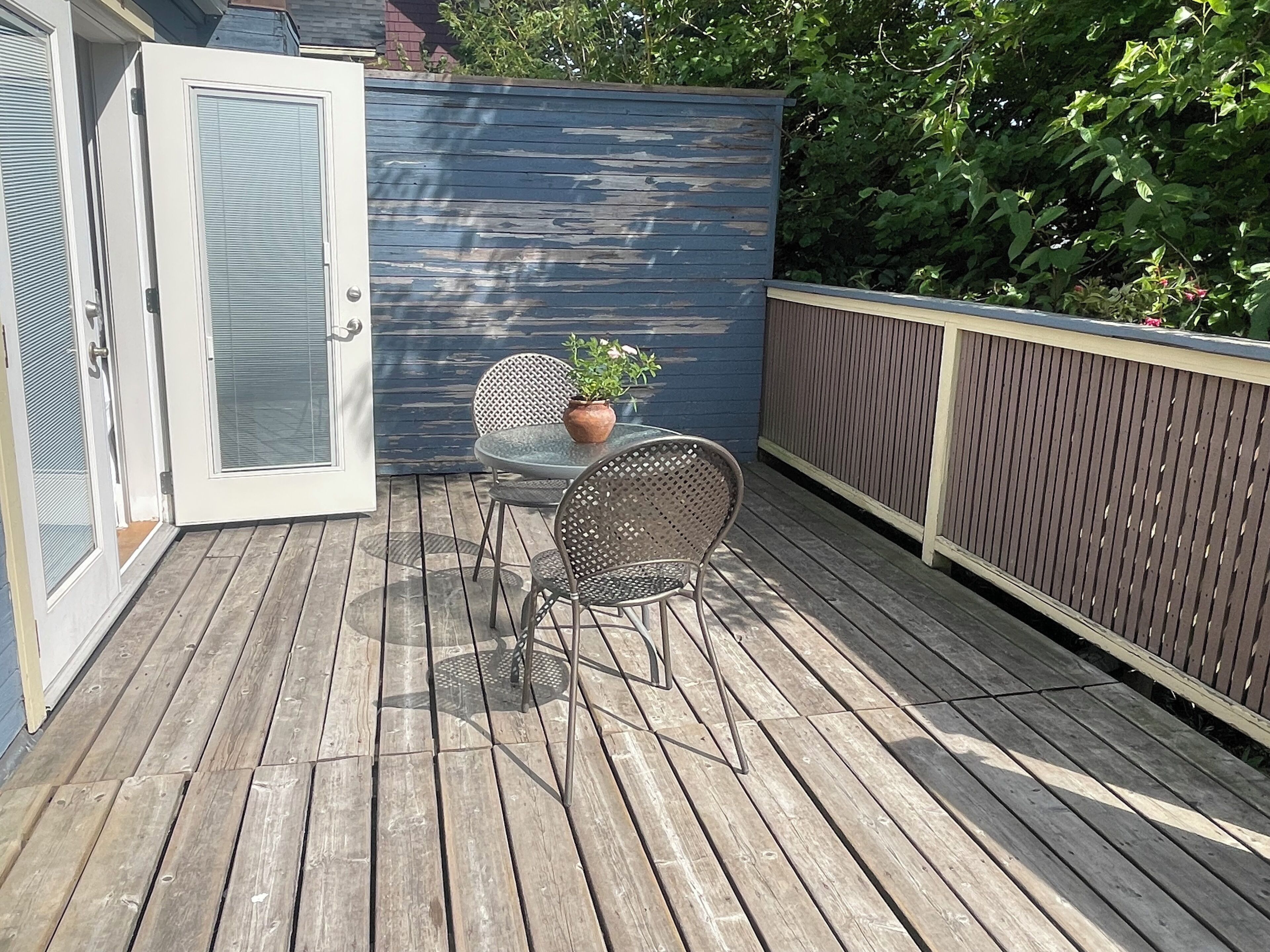 A wooden deck with a table and chairs, a potted plant, and a blue wall with a window.