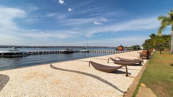 A waterfront promenade with a paved walkway, lounge chairs, and a dock with boats.
