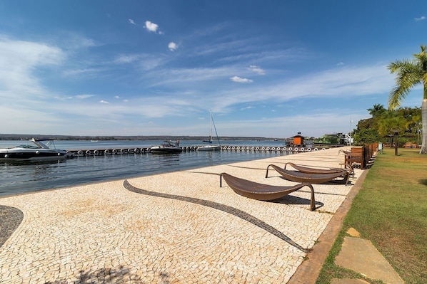 A waterfront promenade with a paved walkway, lounge chairs, and a dock with boats.