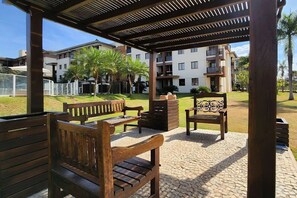 A covered outdoor seating area with wooden benches and a decorative chair, surrounded by a paved walkway and greenery.