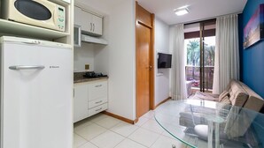 Une cuisine compacte équipée d’un four à micro-ondes, d’une cuisinière et d’un réfrigérateur. On y trouve une table en verre avec des chaises et un canapé dans un espace de vie donnant sur des palmiers.
