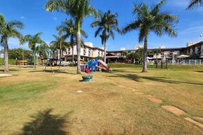 A playground with slides and swings, surrounded by palm trees and a building in the background. - Apt Facing Lake Paranoa - Clr314 (Brasília)
