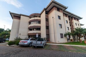 Un edificio de varios pisos con un diseño moderno, que presenta una fachada curva y un techo plano. Hay dos coches aparcados al frente, y el edificio está rodeado de vegetación y palmeras.