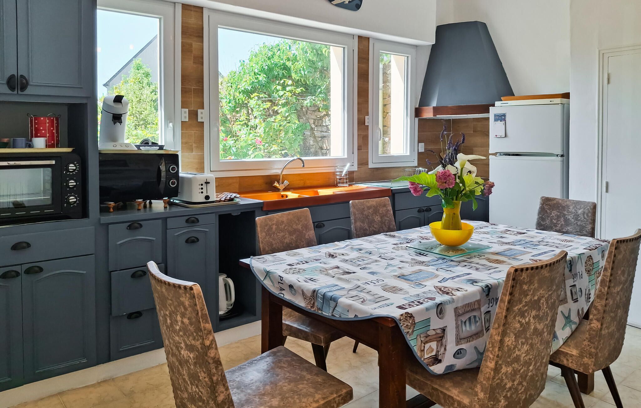Une cuisine avec des meubles de couleur grise, un four encastré, un grille-pain et un évier. On y trouve une table à manger avec un vase jaune et des chaises.