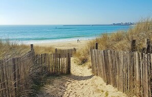 En sti langs en sandstrand, der fører hen mod havet, med et træværk hegn på den ene side og frit udsyn til havet og bygninger i det fjerne.