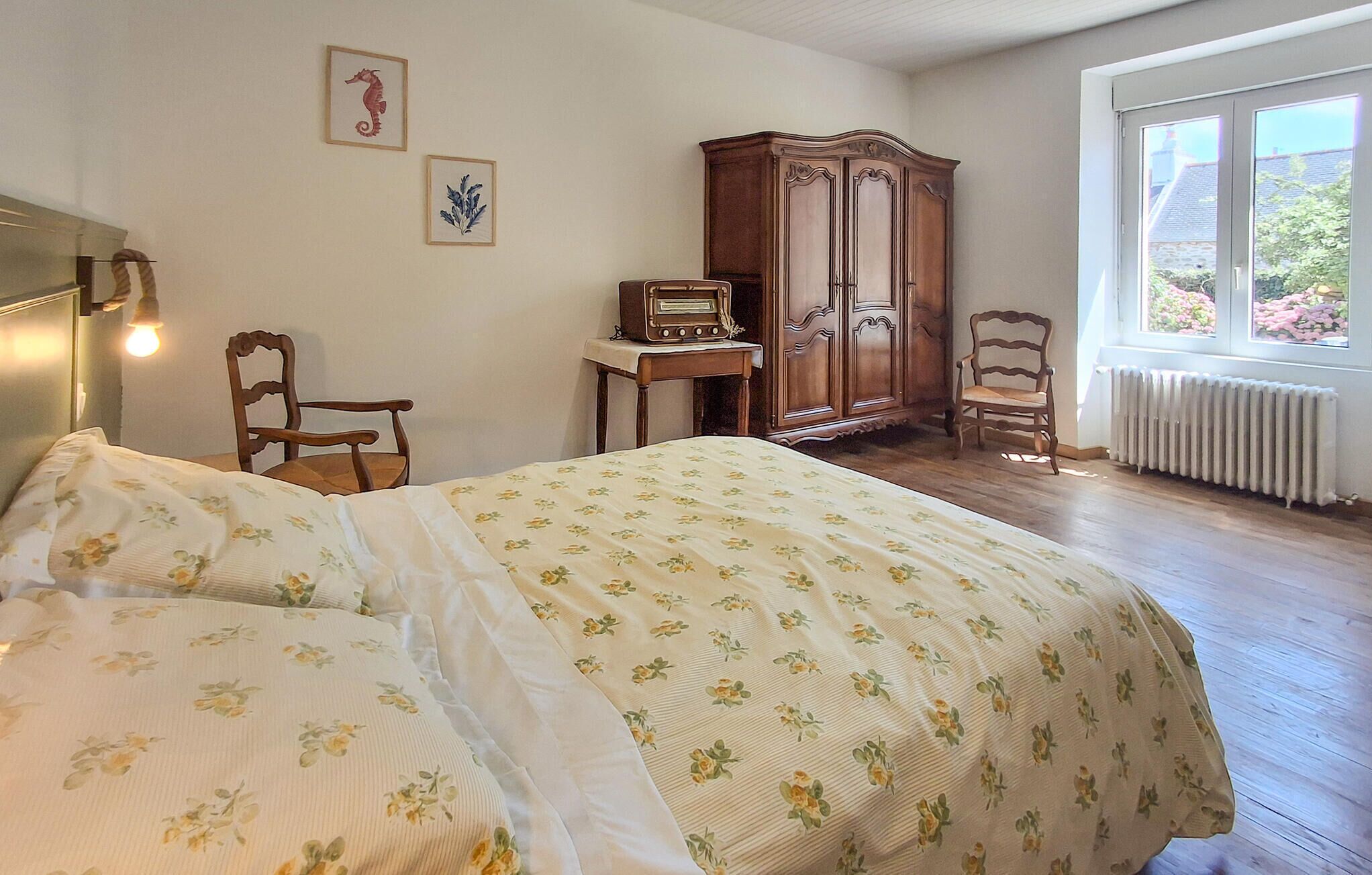 A bedroom with a bed, two chairs, a wooden cabinet, a vintage radio, and a window with a view of greenery.