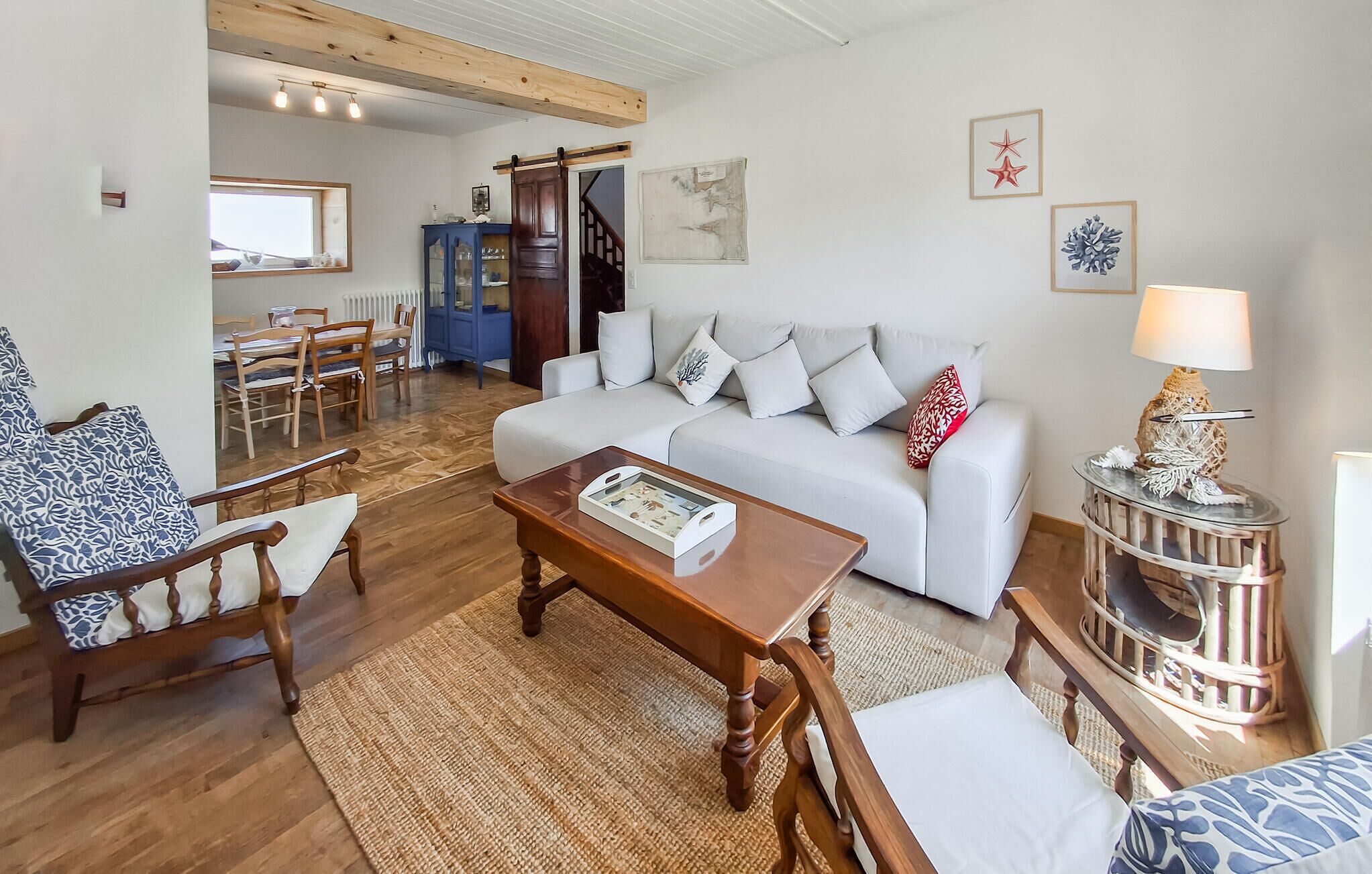 Ein Wohnzimmer mit einem weißen Sofa, einem Couchtisch aus Holz, einem gemusterten Sessel und einem kleinen Beistelltisch mit einer Lampe. Im Hintergrund ist eine Essecke mit Holztisch und Stühlen zu sehen.