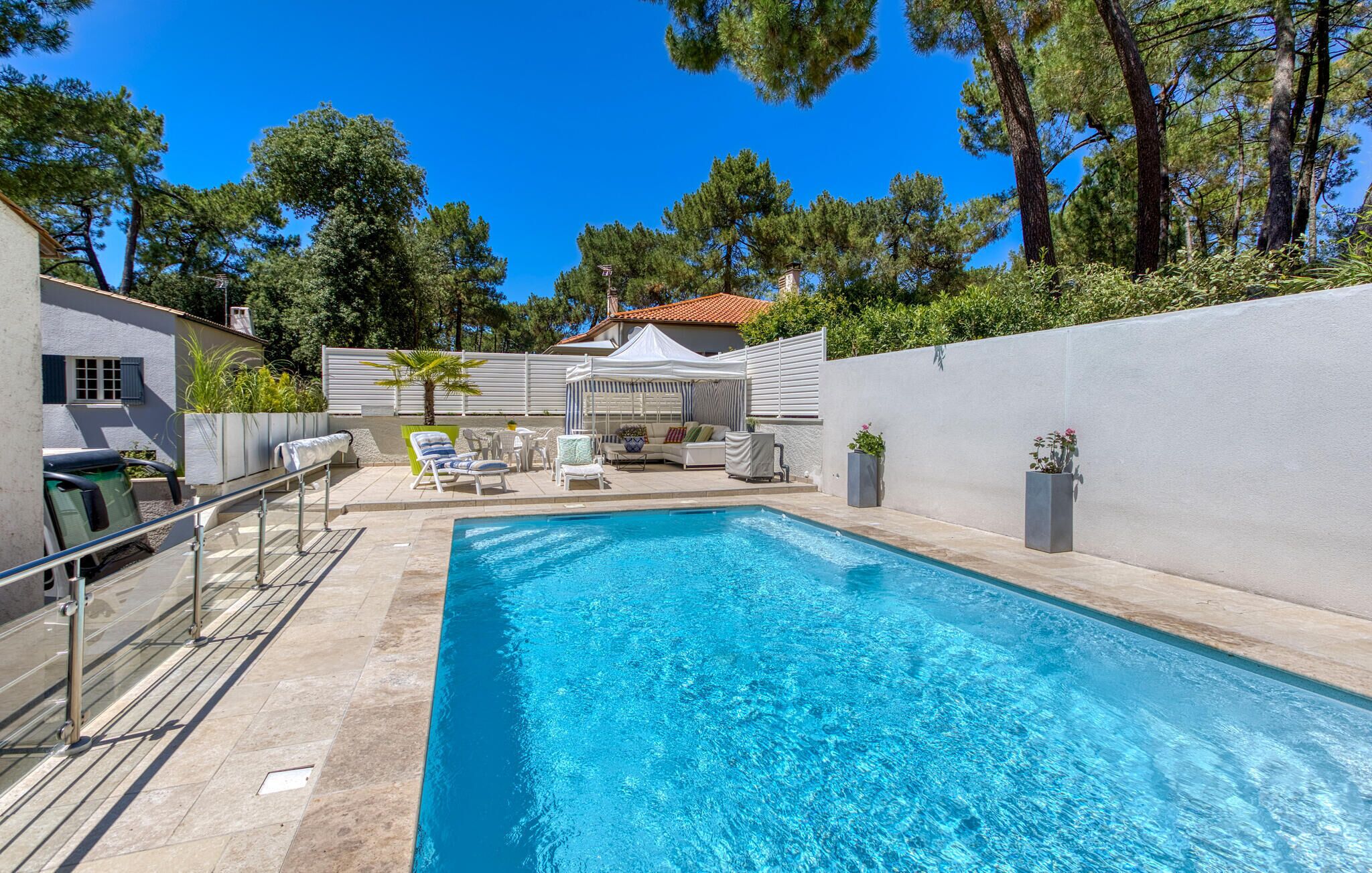 A swimming pool with lounge chairs and a covered seating area, surrounded by a white wall and trees.