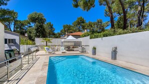 Una piscina con lettini e una zona relax coperta, circondata da un muro bianco e alberi.