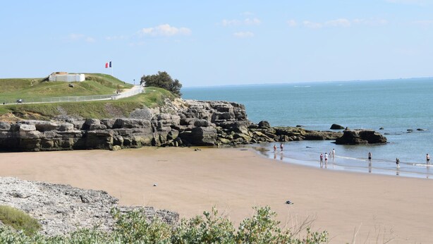 Una spiaggia costiera con scogliere rocciose, un pendio erboso con un edificio e un cielo azzurro limpido.