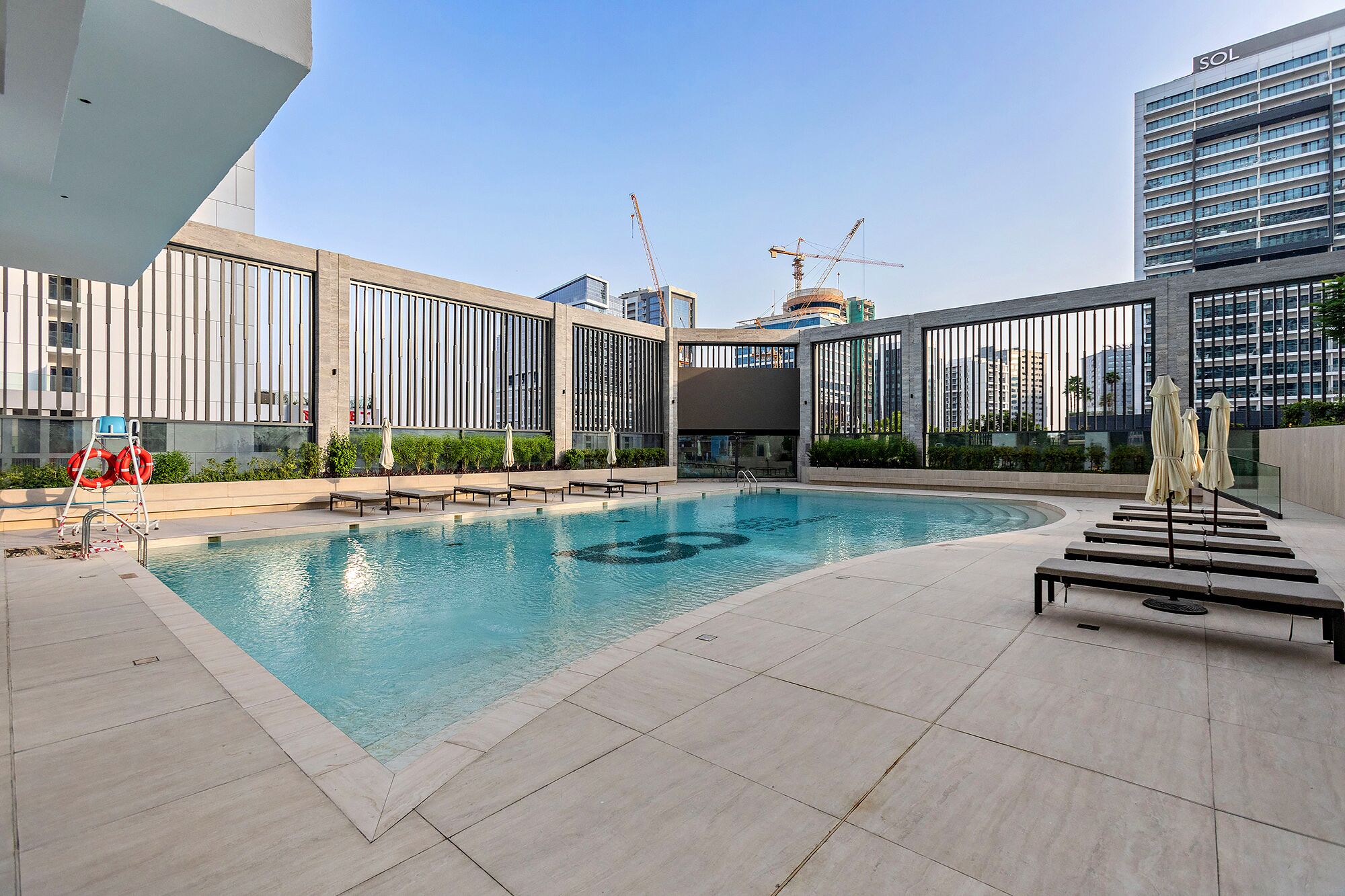 Una piscina en la azotea con sillones de descanso y sombrillas, rodeada de edificios modernos y grúas de construcción.