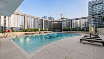 Una piscina en la azotea con sillones de descanso y sombrillas, rodeada de edificios modernos y grúas de construcción.