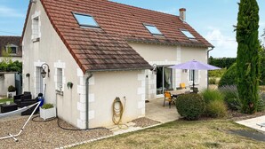 A two-story house with a sloped roof, a small patio with outdoor furniture, and a garden area. - Gite avec piscine et jacuzzi (Seigy)