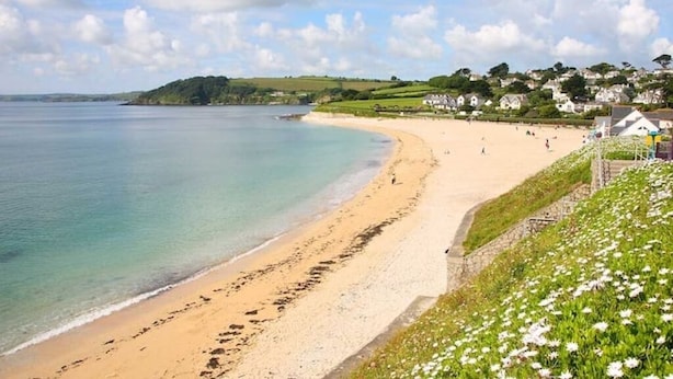 A coastal beach with a sandy shore, a clear blue sea, and a backdrop of houses on a green hillside.