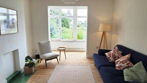 A living room with a dark blue sofa, a white armchair, a small round table, a floor lamp, and a large window with a view of greenery.